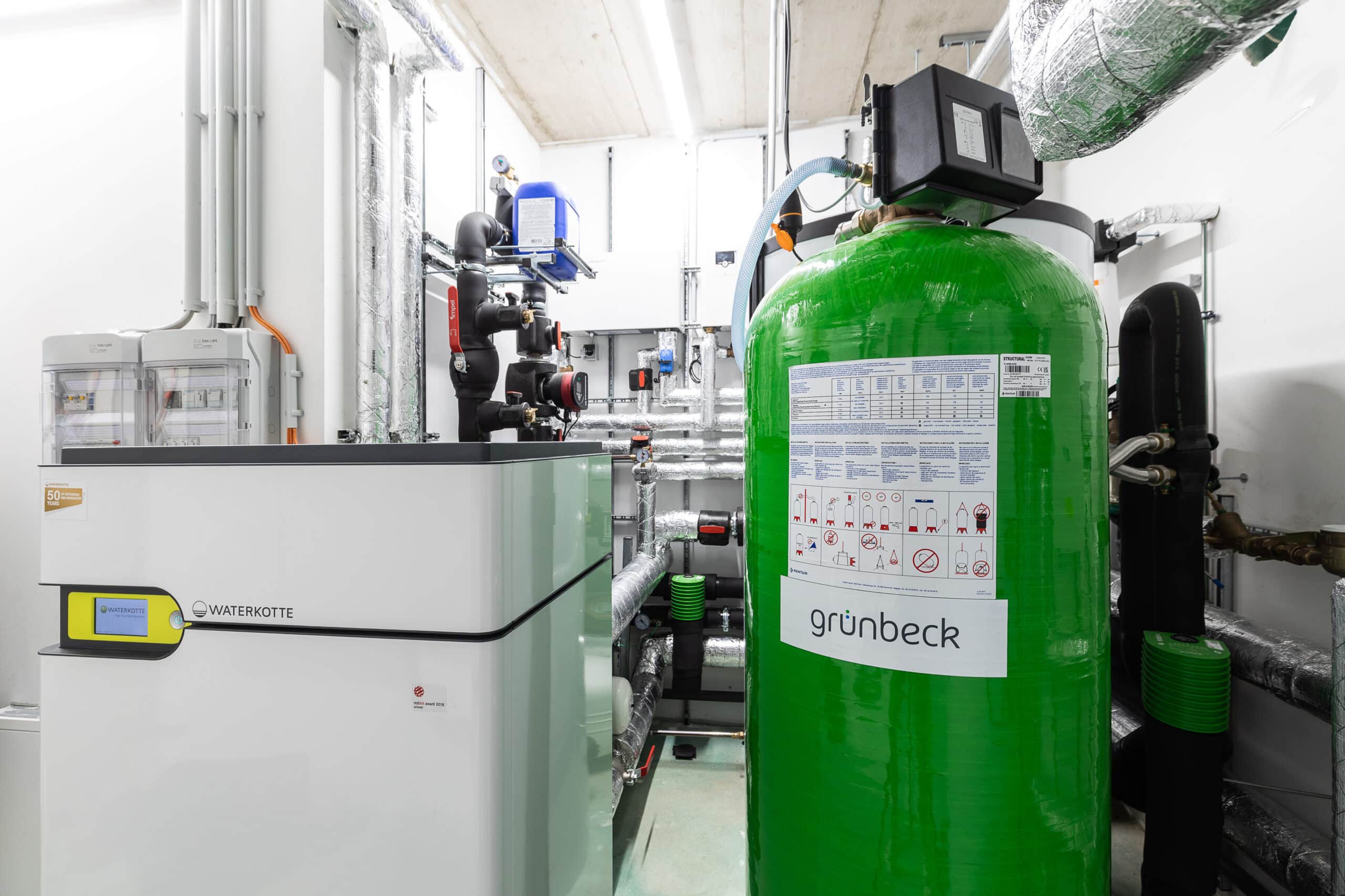 Grüne Wasseraufbereitungsanlage neben einer Wärmepumpen-Inneneinheit und gedämmten Rohrleitungen